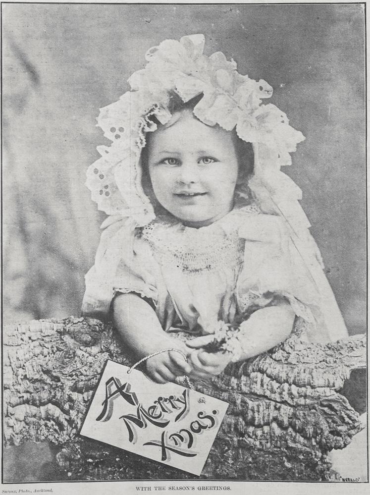 portrait of a very young girl wearing a frilly bonnet and dress, leaning on a log and holding a sign 'A Merry Xmas'